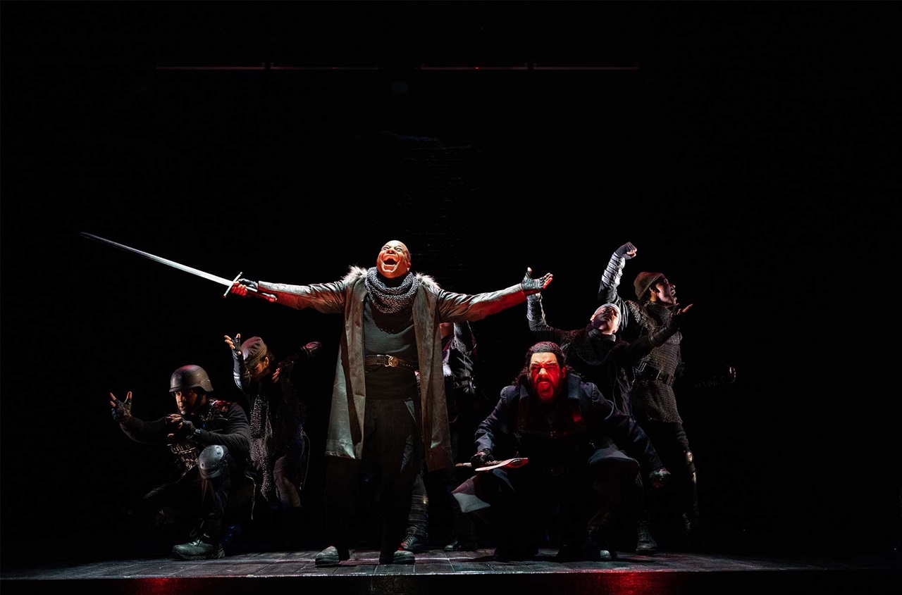 A man standing in the middle of the stage holding a sword with solders all around him.