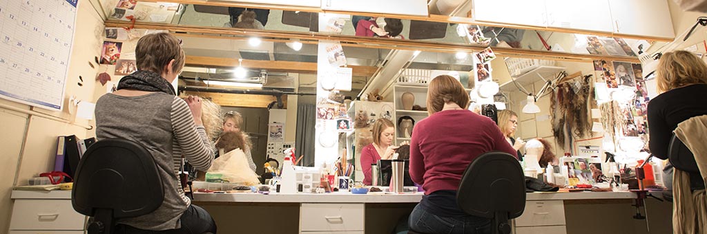 Three people sitting in chairs facing a large mirror working on wigs.