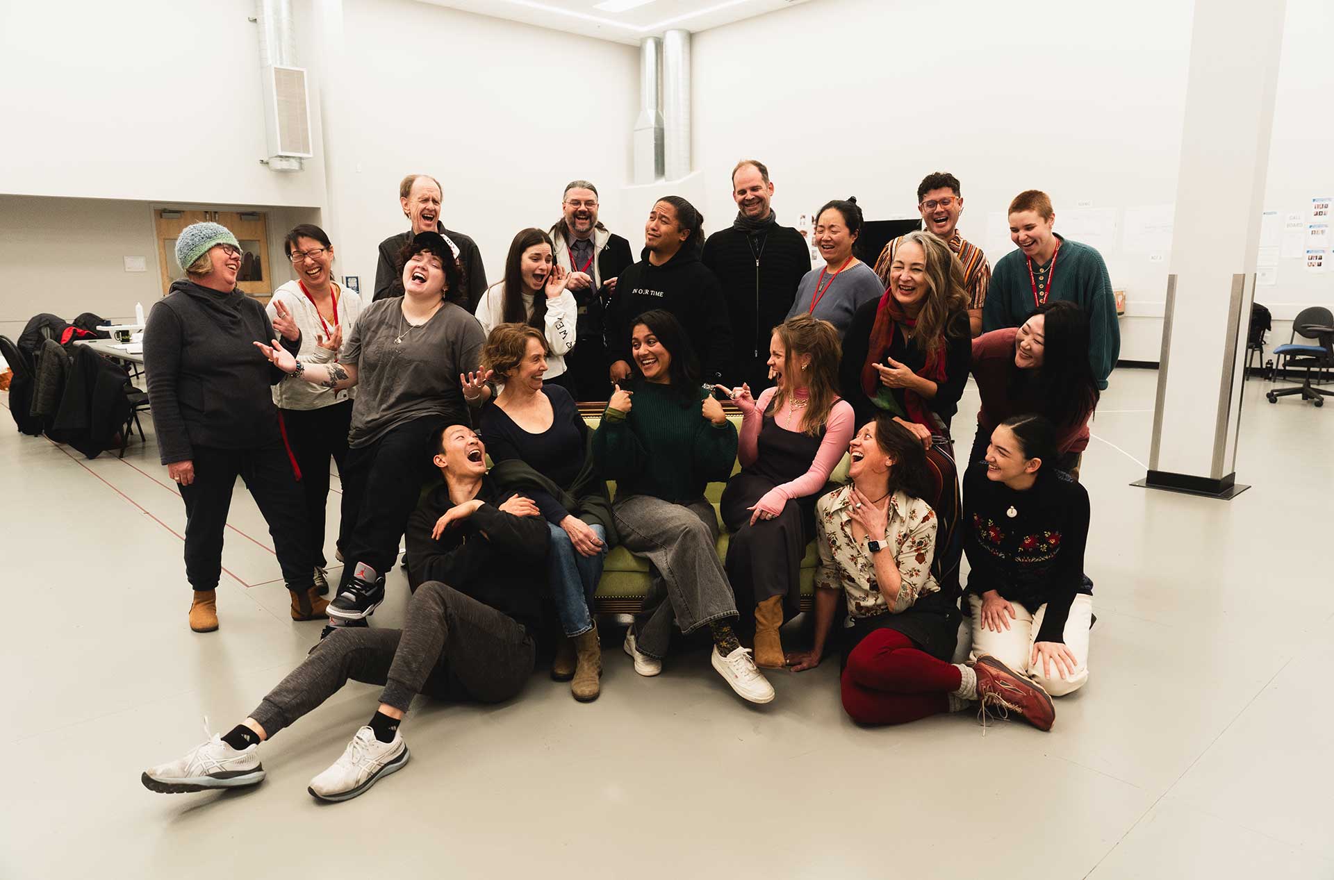 A joyful group portrait of the rehearsal team, with everyone laughing and posing playfully. The group stands and sits together in a bright rehearsal space, conveying camaraderie and energy.