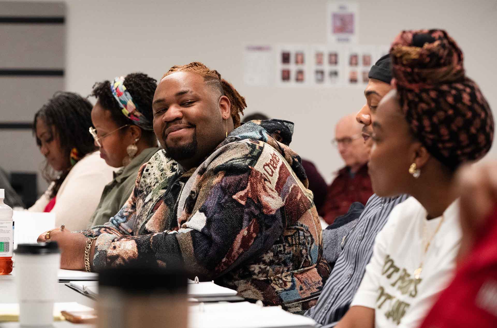 A person in a patterned sweater smiles at the camera while sitting at a long table. Others around them are focused on their work or conversation.