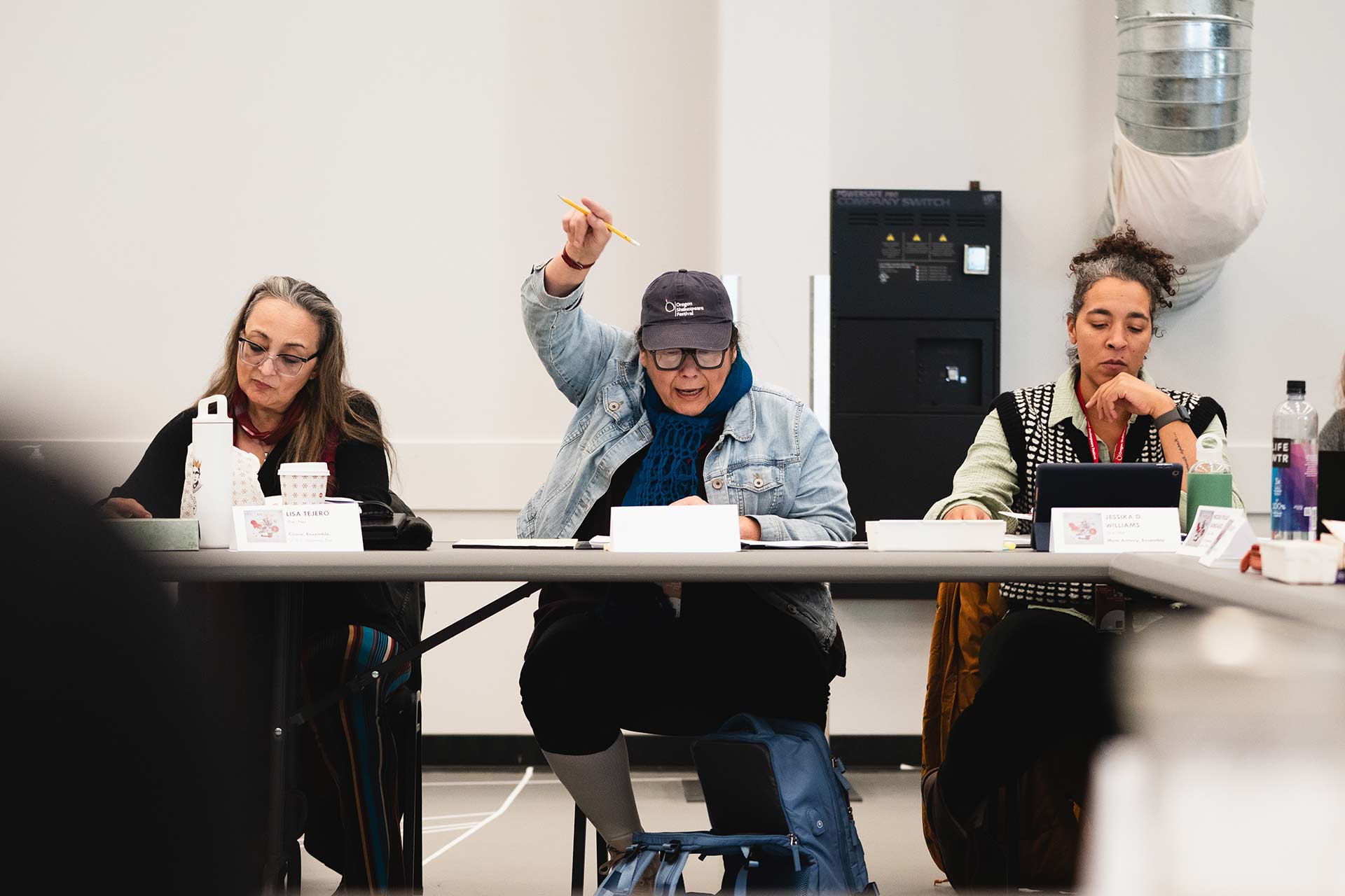 A team member raises a hand during a rehearsal. The person is wearing a denim jacket, hat, and scarf. Two others beside them are focused on their scripts and notes.