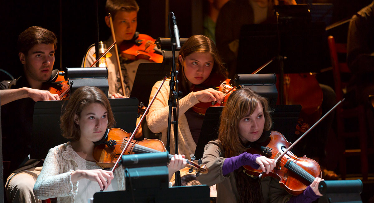 A close-up of musicians performing in OSF's 2014 production of Into the Woods. The image focuses on violin players seated in the orchestra, their expressions intense and focused under warm stage lighting.